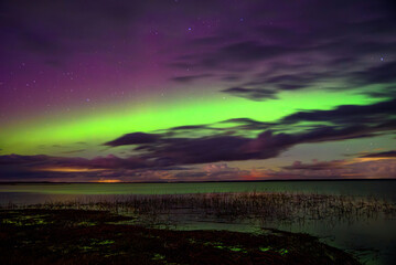 Obraz premium Vivid northern lights glow in green and purple hues over a calm lake beneath a starry night sky with clouds.