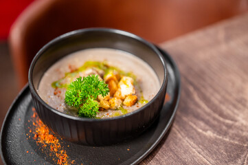 Creamy mushroom soup with croutons and fresh parsley in a black bowl on a rustic wooden table, garnished with herb oil and paprika powder