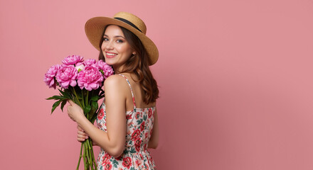 Smiling young woman in summer hat holding bouquet of pink peonies. Happy girl in floral dress posing on pink background with copy space
