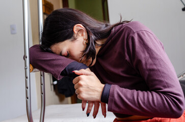 Depressed disabled young woman with her head resting on a crutch sitting on a couch.Sad woman feeling pain.