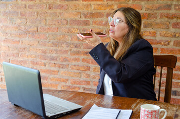 Mature woman working with a laptop and sending an audio text with her mobile phone.Woman taking notes while talking on mobile phone.