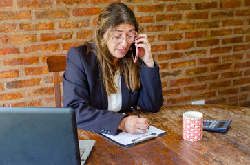 Mature woman working with a laptop and having a conversation on phone.Woman taking notes while talking on mobile phone.