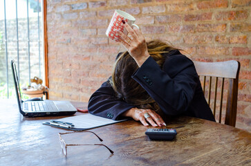 Exhausted mature female employee feeling fatigue lying on table and lifting cup of coffee lack of energy in morning office tired of stress problems sleeping at workplace overwork concept
