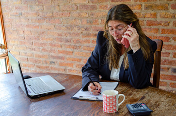 Mature woman working with a laptop and having a conversation on phone.Woman taking notes while talking on mobile phone.