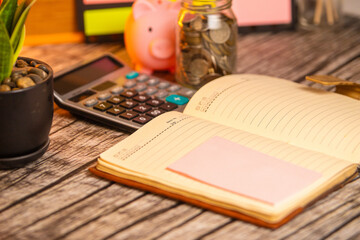 Image of an open notebook with a pink sticky note is in focus on a wooden table, with a calculator, a potted plant, a piggy bank, and a jar of coins slightly blurry in the background.  