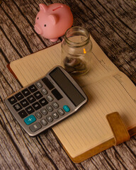 image shows small pink piggybank, a partially filled glass jar of coins, and a calculator resting on an open notebook. The image suggests budgeting, saving, and financial planning on a wooden surface.