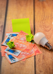Image of a $50 barbados dollar banknotes, a yellow sticky note, a small LED lightbulb, and green crumpled paper are on a wooden surface, suggesting energy saving and financial management.