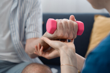 Female nurse assisting elderly man with hand exercise using 1kg dumbbell at home, highlighting rehabilitation, strength training, and senior care therapy in a home healthcare setting.