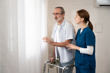 Obraz premium Elderly man with a walker standing by the window, assisted by a caregiver. Concept of home care, recovery, rehabilitation, or senior support.