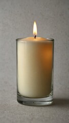 Lit white candle in clear glass holder on neutral background
