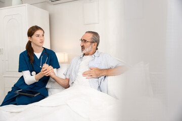 Obraz premium Young female nurse supporting senior male patient in bed during home care visit. Ideal for healthcare, elderly support, nursing, recovery, medical assistance, and caregiving visuals.