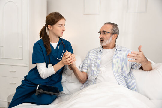 Young female nurse supporting senior male patient in bed during home care visit. Ideal for healthcare, elderly support, nursing, recovery, medical assistance, and caregiving visuals.