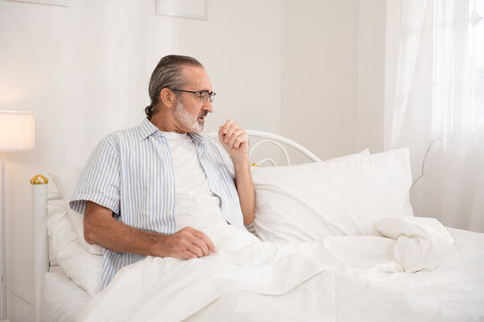 Senior man sitting in bed and coughing, showing signs of illness or respiratory discomfort in bright bedroom. Great for healthcare, cold, flu, elderly wellness, and medical awareness visuals.