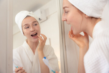 Young woman in white bathrobe and towel checking her teeth in mirror with toothbrush in hand. Morning dental care routine, oral hygiene, healthy lifestyle, brushing, teeth, dental, oral, hygiene,