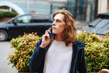 Young man talking on the phone while sitting near a green bush in an urban setting