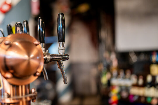 A close-up, selective focus shot of a row of shiny chrome and copper beer taps on a bar counter with a blurred background of a pub.