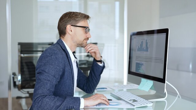 Businessman in a blue jacket analyzing financial data charts on computer, making important business decisions in modern office. Business people concept