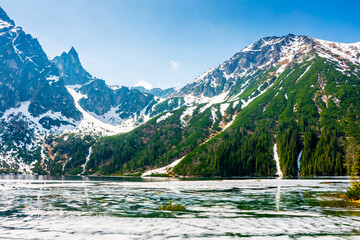 mountain lake in tatra national park. morskie oko surrounded with rugged snow capped peaks in early spring. beautiful view of reflection and ice on water. green environment background under blue sky © Lesia