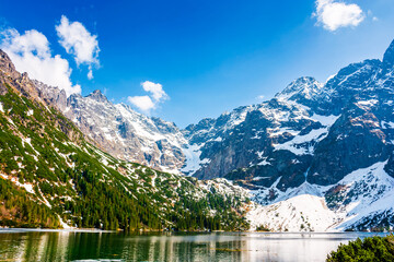 mountain lake in tatra national park. morskie oko surrounded with rugged snow capped peaks in early spring. beautiful view of reflection and ice on water. green environment background under blue sky © Lesia