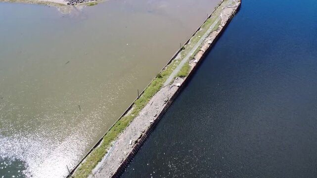 Aerial view of the remains of an old abandoned wharf separating two bodies of water of different colors, blue and brown, with the reflection of the sun sparkling on the waves. Campbellton, NB, Canada,