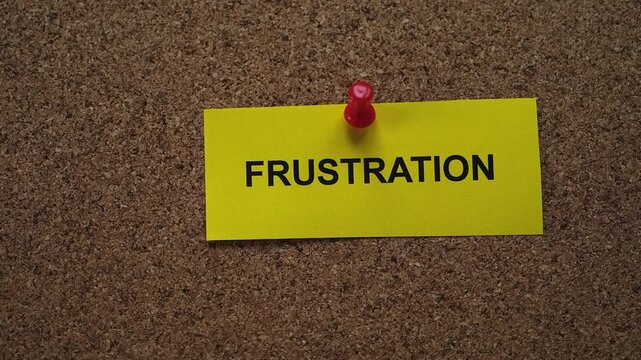 A yellow paper note with the word Frustration on it pinned to a cork board. Close up.