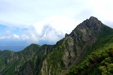 Serrated rocky summit of Mount Ashibetsu, Hokkaido, towers above alpine shrubs and a scree gully under building cumulus clouds and a bright summer sky.