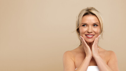 Fototapeta premium Antiage Skincare. Happy Attractive Middle Aged Female Touching Face , Beautiful Woman With Flawless Skin Standing Wrapped In Towel Over beige Studio Background, Copy Space. High quality photo