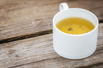 A white cup filled with herbal tea sits on a rustic wooden surface, showing warm tones and small floating herbs in the golden liquid.