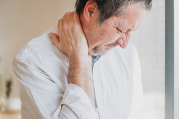 Unshaved businessman suffering from severe neck pain, holding neck in discomfort at office, stressed man feeling tension and stiffness.