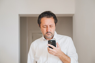 Mature adult unshaved businessman standing in bright modern office, looking at smartphone screen and reading message.