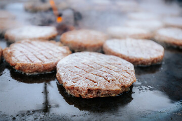 A hand with a ring uses a fork and spatula to flip raw burger patties on a smoky circular grill over glowing charcoal, with flames and steam rising.