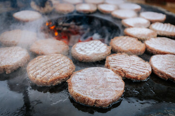 A hand with a ring uses a fork and spatula to flip raw burger patties on a smoky circular grill over glowing charcoal, with flames and steam rising.