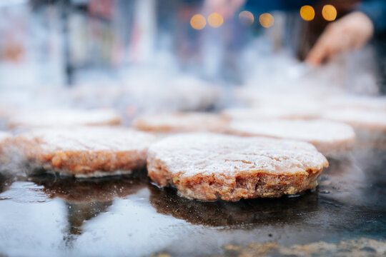 A hand with a ring uses a fork and spatula to flip raw burger patties on a smoky circular grill over glowing charcoal, with flames and steam rising.
