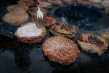 Overhead shot of multiple burger patties cooking on a circular grill surrounding hot charcoal, with a hand flipping one patty using fork and spatula.