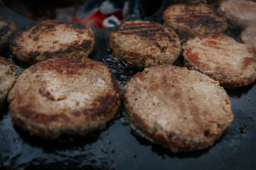 Overhead shot of multiple burger patties cooking on a circular grill surrounding hot charcoal, with a hand flipping one patty using fork and spatula.