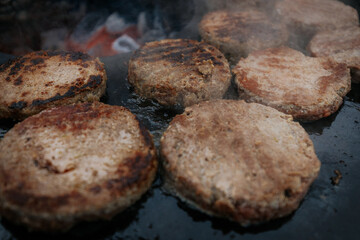 burger patties sizzling on a flat grill above glowing charcoal, with a hand flipping them using fork and spatula. Steam and grill marks visible.