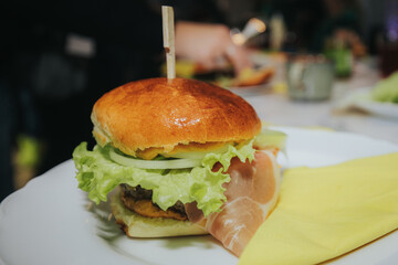 A close-up of a juicy cheeseburger with lettuce, cheddar, onions, and a beef patty in a golden bun, served on a white plate at a lively indoor gathering.