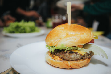 A delicious homemade cheeseburger with lettuce, onion, pickles, tomato, and melted cheddar, served on a soft bun with a wooden skewer on a white plate.
