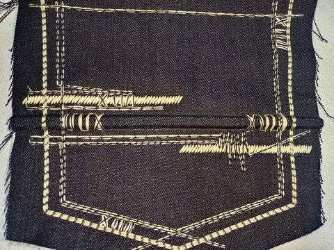 Extreme close-up macro photograph of a dark indigo denim back pocket design. The focus is on the intricate, heavy, contrast stitching in a thick beige or gold thread.