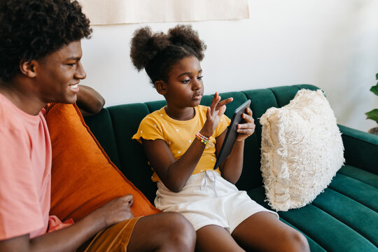 Afro-haired siblings relaxing on sofa, browsing together on tablet in living room