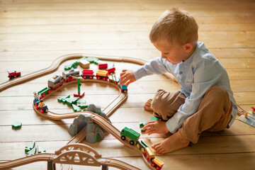 Boy barefoot playing with colourful wooden toy train set, assembling tracks and wagons. Concept of learning through play, creativity, and early childhood development at home. Eco-friendly wooden toys