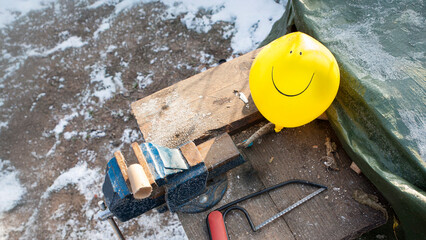 Workbench scene outdoors in winter. Yellow balloon with smiley face next to vise, saw and wood shavings. Work life balance. Positivity at work and mental health. Humor and anti-stress