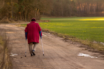 Elderly woman with crutches walking outdoors toward wooded area outdoors. Physical activity for seniors. Rural healthcare and accessibility. Loneliness in old age. Healthy lifestyle in senior years