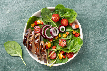 Top view of a healthy chicken salad with spinach leaves, cherry tomatoes, red onion, and colorful bell peppers on a green textured background.
