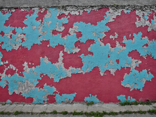 ﻿Verfall trifft Natur: Rote und blaue Abblätterung auf Beton, während Gras und Moos die einstige Wand erobern – ein urbanes, melancholisches Bild von Zeit, Verlorenheit und Erneuerung.