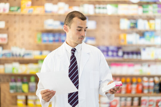 Positive young male pharmacist standing in chemist's shop holding box with medicament and a sheet of recipe