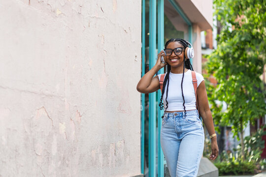 Smiling Latina woman with braided hair listening to music on the street while wearing headphones. 