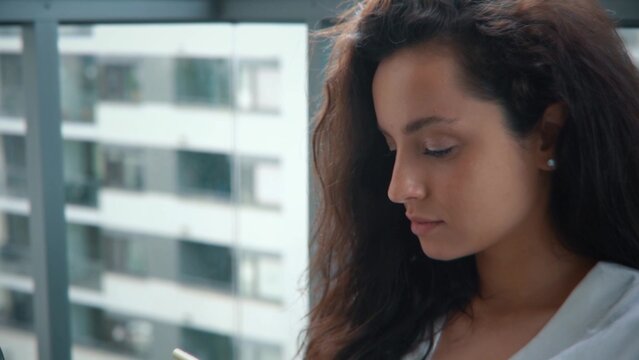 Camera moving back from happy young housewife sitting on balcony in apartment texting on smartphone and smiling. Caucasian attractive brunette woman in white robe with laptop browsing on cellphone.