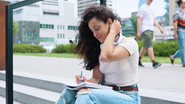 Caucasian young beautiful female student sitting on stairs outdoors near college or university and writing in her notebook. Pretty brunette writes in copybook in street. Studying. Education concept. - Powered by Adobe