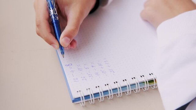 Hand writing math equations in dotted notebook with blue pen, solving arithmetic problems including negative numbers, educational activity, student homework, learning study session, school concept